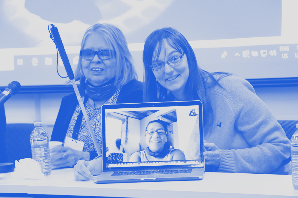 two people sitting at a conference table that has a laptop opened and another person on the screen
